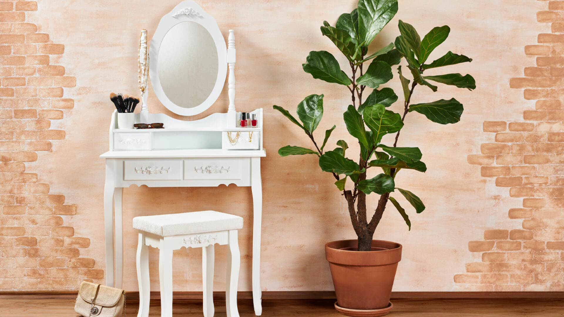 Ein weißer Vintage-Schminktisch mit Spiegel und passendem Hocker steht neben einer großen Topfpflanze vor einer Wand mit Ziegelmuster. Make-up-Artikel und Schmuck liegen auf dem Schminktisch. Eine kleine beige Tasche liegt auf dem Boden.