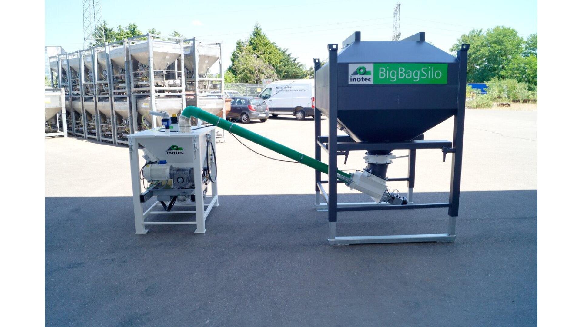 A large gray and green industrial BigBagSilo and a smaller white feed machine are connected by a green tube, set outdoors on a paved area with parked cars and metal racks in the background.
