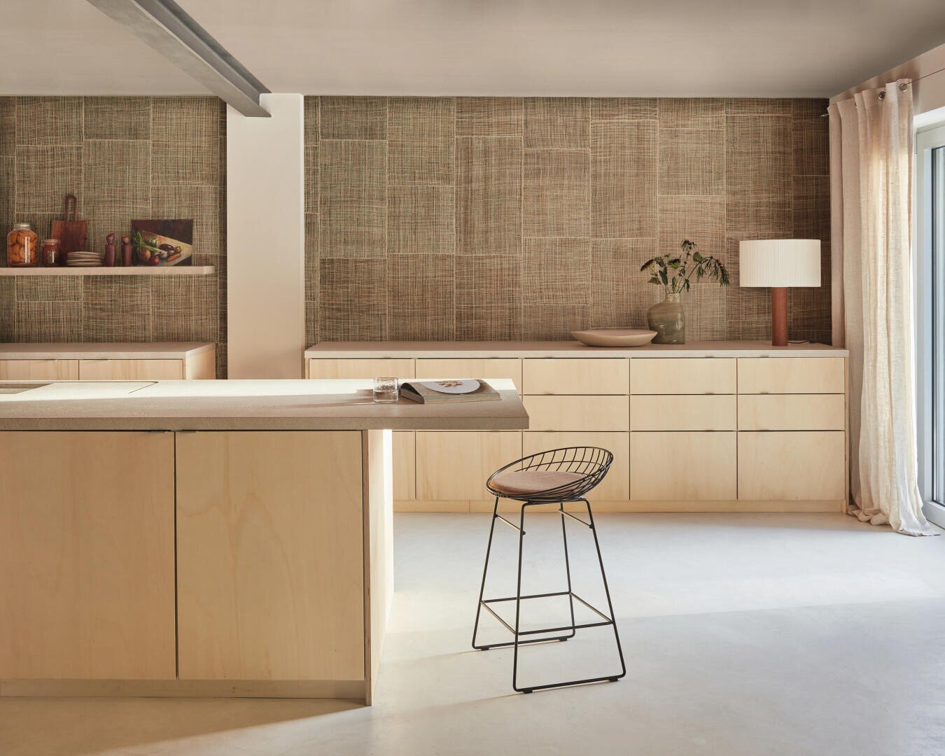 Minimalistische Küche mit hellen Holzschränken, beigefarbener Küchenrückwand, einer Kücheninsel, einem Hocker mit Drahtgestell und Sonnenlicht, das durch bodenlange Vorhänge an einer Glasschiebetür fällt. Eine Lampe und eine Vase schmücken die Arbeitsfläche.