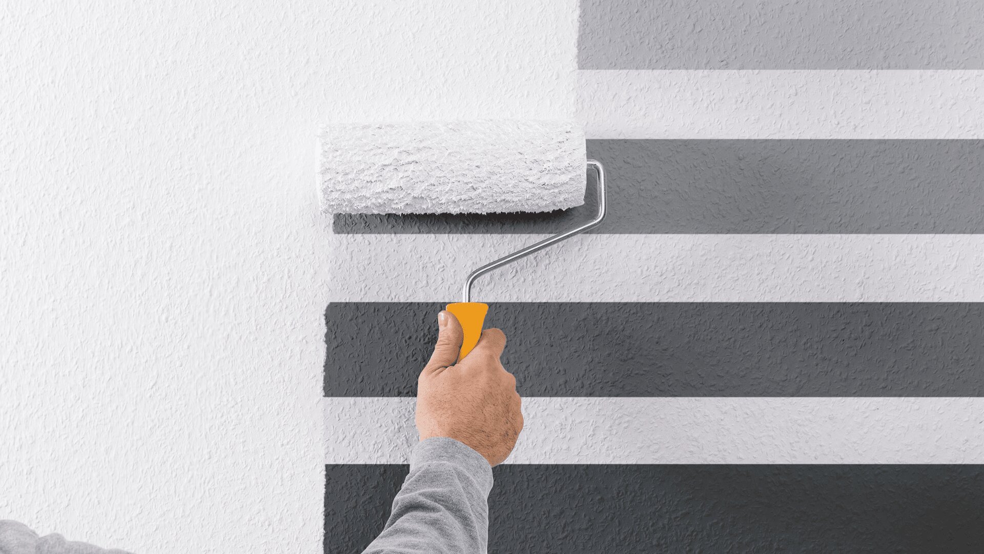 Eine Hand hält eine Farbrolle mit gelbem Griff und überstreicht eine Wand mit horizontalen schwarzen und grauen Streifen und überdeckt sie mit weißer Farbe.