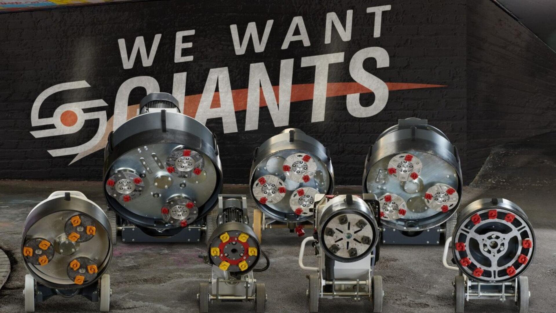 Seven large, industrial pipe inspection robots with visible wheels and cameras are lined up in front of a mural that reads WE WANT GIANTS on a dark brick wall.