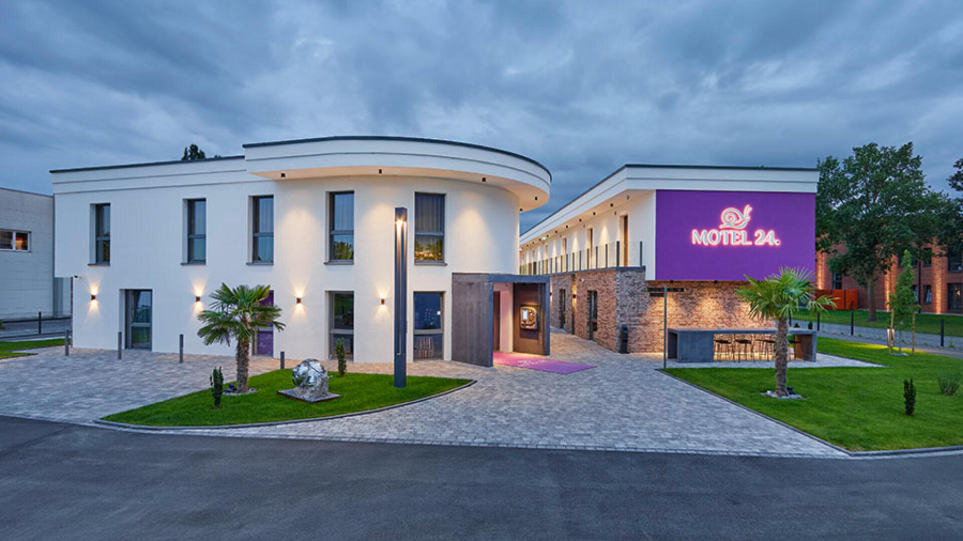 Ein modernes, zweistöckiges weißes Gebäude mit großen Fenstern und einem leuchtend violetten Motel 24-Schild in der Abenddämmerung, umgeben von Palmen, Straßenlaternen und einer gepflasterten Auffahrt.