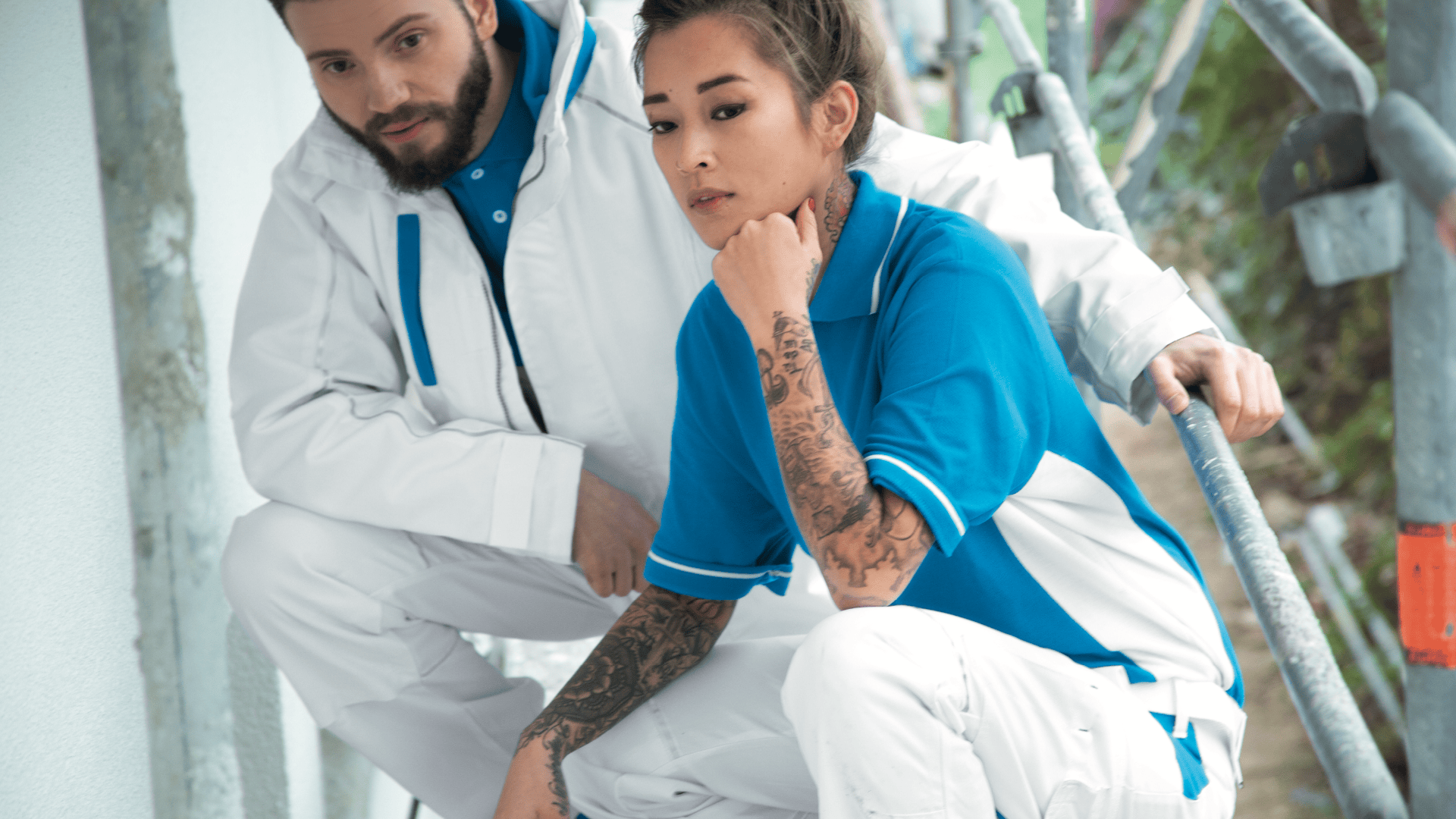 Zwei Bauarbeiter, ein Mann und eine Frau mit Tätowierungen, in blau-weißen Uniformen hocken auf einem Gerüst und blicken ernst in die Kamera.