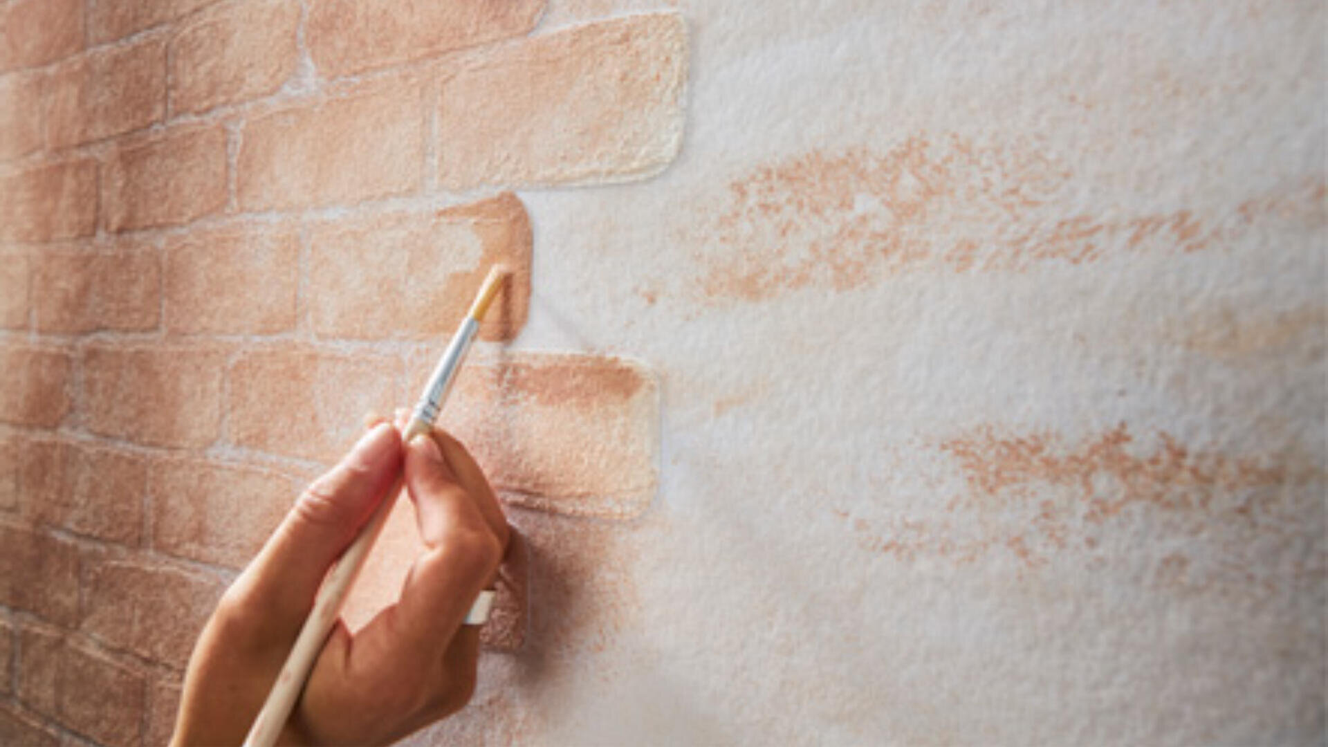 Eine Hand malt mit einem kleinen Pinsel ein Ziegelmuster auf eine weiße Wand und fügt vorsichtig rotbraune Farbe hinzu, um einen realistischen Ziegeleffekt zu erzeugen.