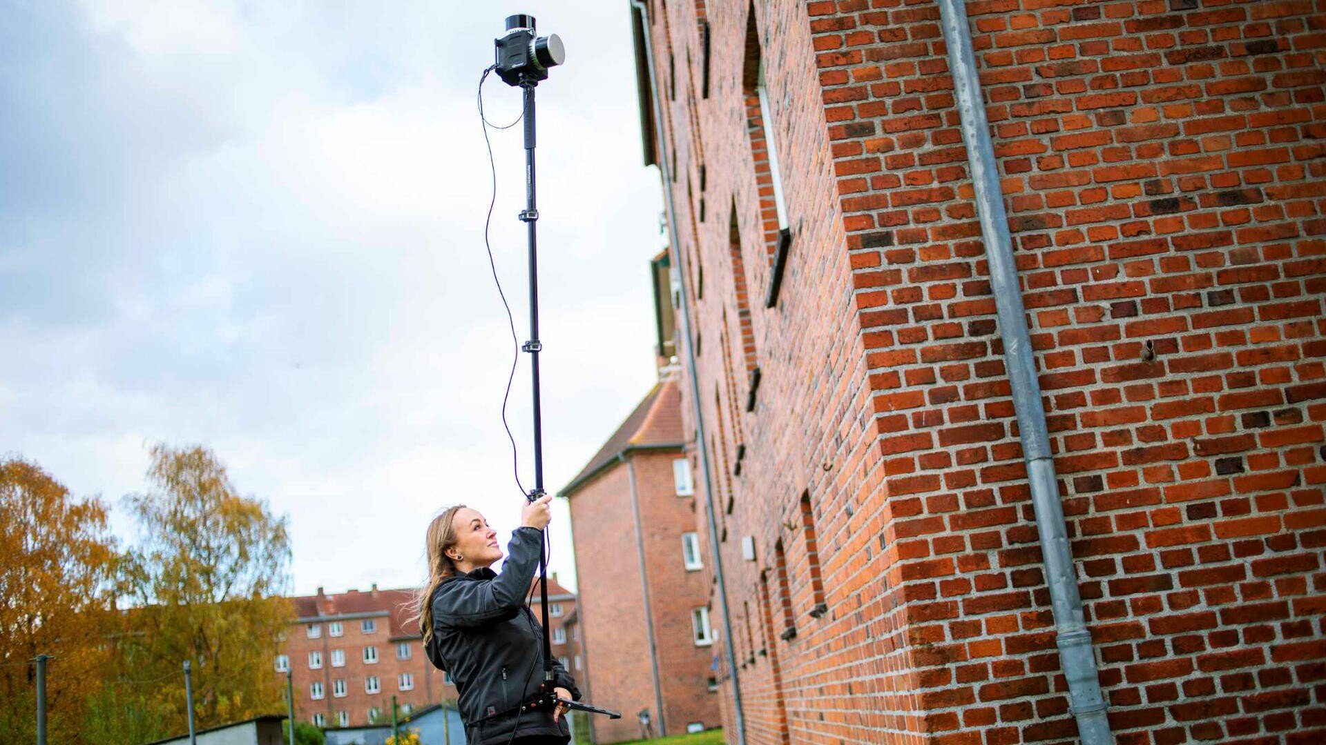 Eine Person im Freien hebt eine Kamera mit einer langen Stange hoch über ihren Kopf und richtet sie auf ein hohes Backsteingebäude. Der Himmel ist teilweise bewölkt, im Hintergrund sind Bäume zu sehen.