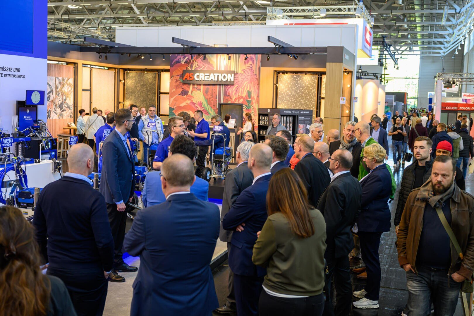 Eine Gruppe von Menschen versammelt sich an einem belebten Messestand mit Maschinenausstellungen. Die Besucher, meist in Businesskleidung, verfolgen eine Präsentation. Stände, Schilder und helle Lichter füllen den Hintergrund.