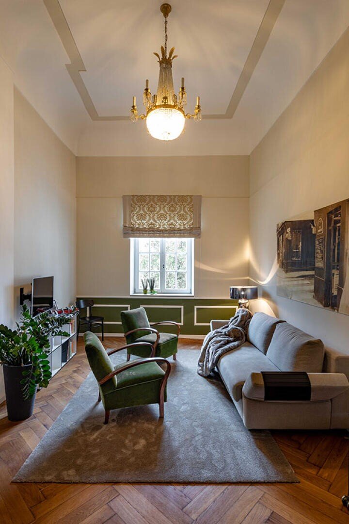 Ein gemütliches Wohnzimmer mit einem grauen Sofa, zwei grünen Sesseln, einem Teppich, einem Fernseher auf einem Ständer, einer großen Zimmerpflanze und einem Fenster, das natürliches Licht hereinlässt. Von der hohen Decke darüber hängt ein Kronleuchter.