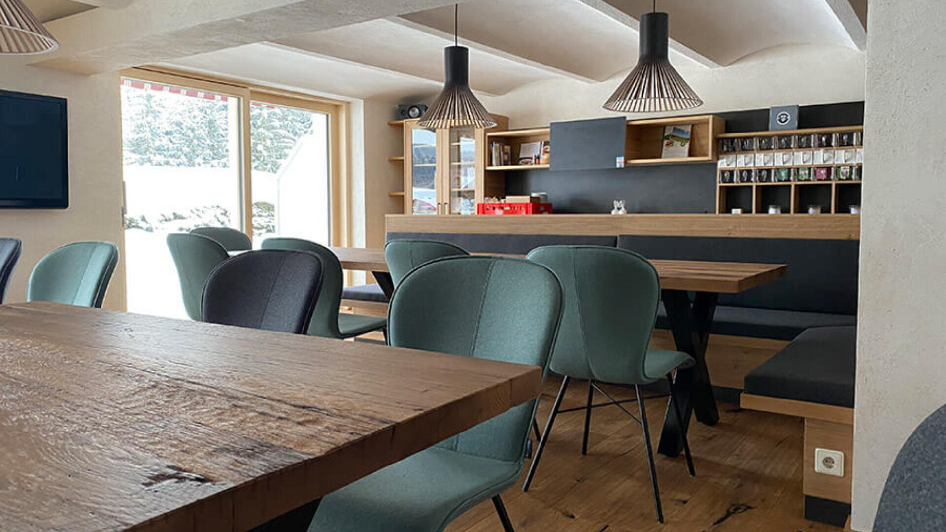 Ein gemütliches, modernes Café-Interieur mit Holztischen, gepolsterten Stühlen, Hängelampen, Regalen mit Gläsern und einem Fenster, das natürliches Licht hereinlässt. Draußen ist eine verschneite Landschaft zu sehen.