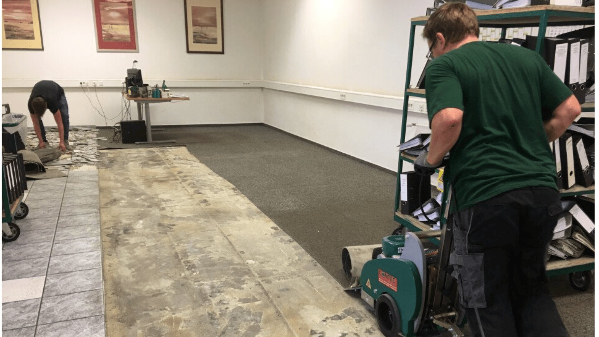 Two people removing old flooring in an office; one is using a floor stripping machine while the other gathers debris. The room has shelves with files and framed pictures on the wall.