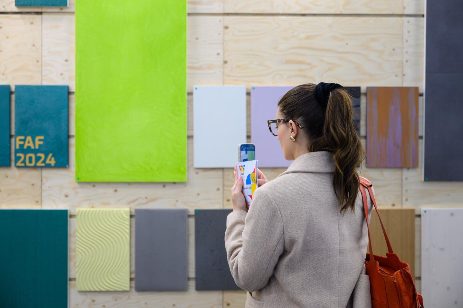 Eine Frau mit Pferdeschwanz und Brille fotografiert bunte, strukturierte Paneele an einer Wand. Sie hält ein Telefon und eine Farbmusterkarte in der Hand und trägt eine orangefarbene Tasche über der Schulter. Auf einem Paneel steht „FAF 2024“.