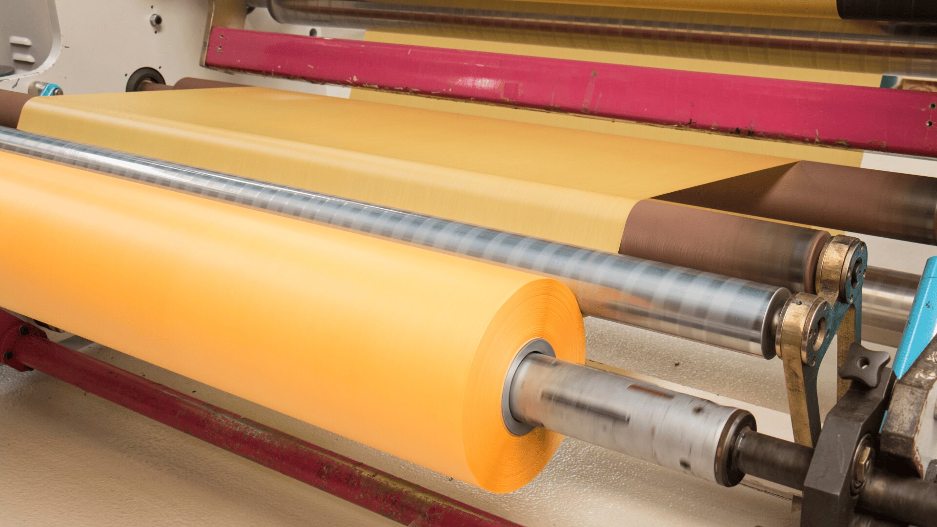 A close-up of a machine unwinding a large roll of yellow paper or film onto rollers in an industrial setting, with visible metal rods and pink structural supports.