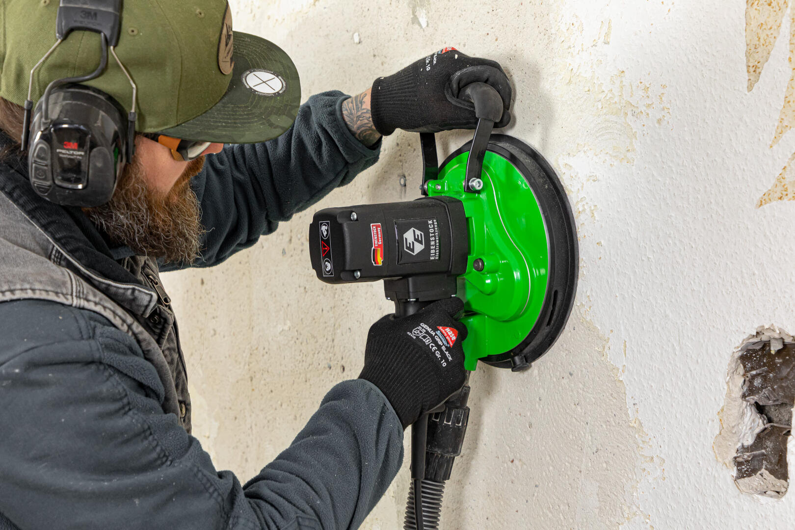 Ein Mann in Schutzkleidung schleift oder glättet mit einem grünen Elektrowerkzeug eine weiße Wand. Er trägt Handschuhe, eine Mütze und einen Kopfhörer.
