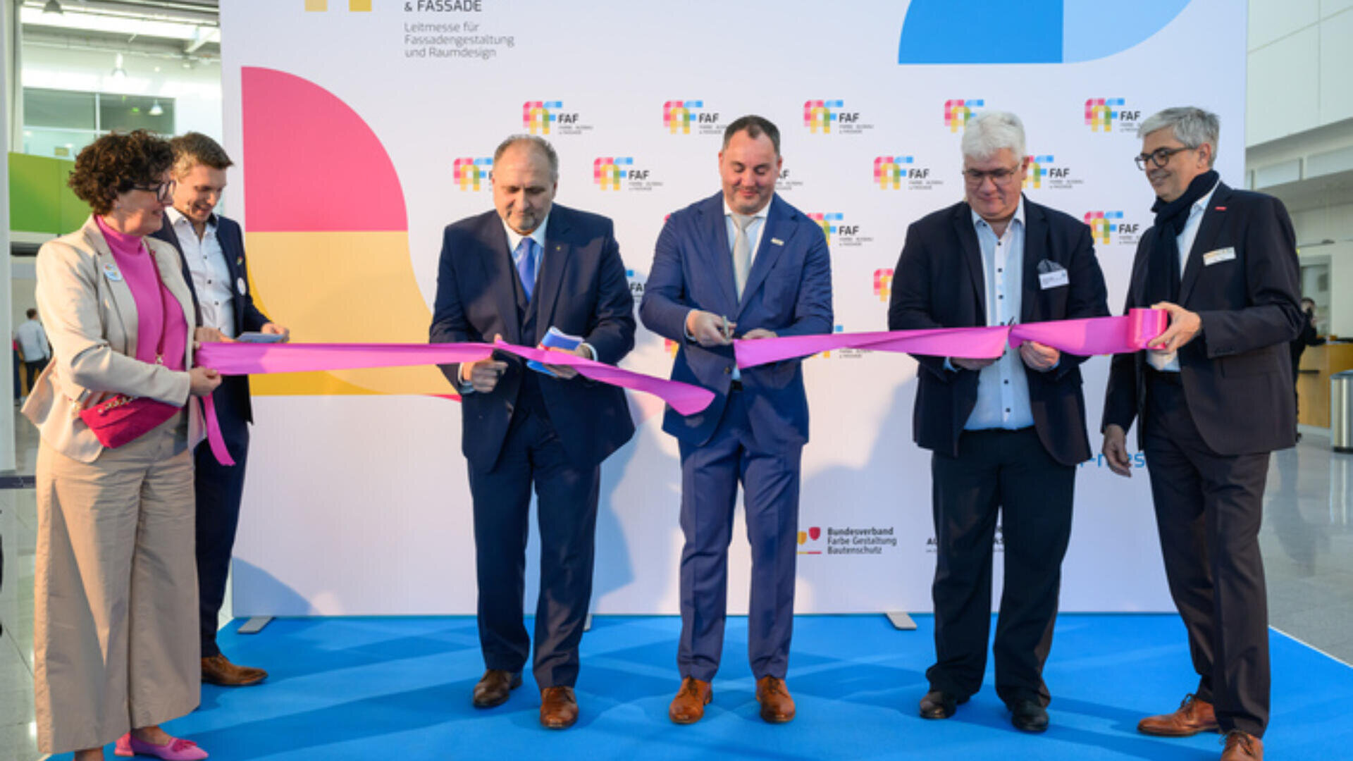 Sechs Personen in Geschäftskleidung zerschnitten bei einer Indoor-Veranstaltung ein langes rosa Band und standen dabei auf einem blauen Teppich vor einem Markenhintergrund, was eine feierliche Eröffnung symbolisierte.