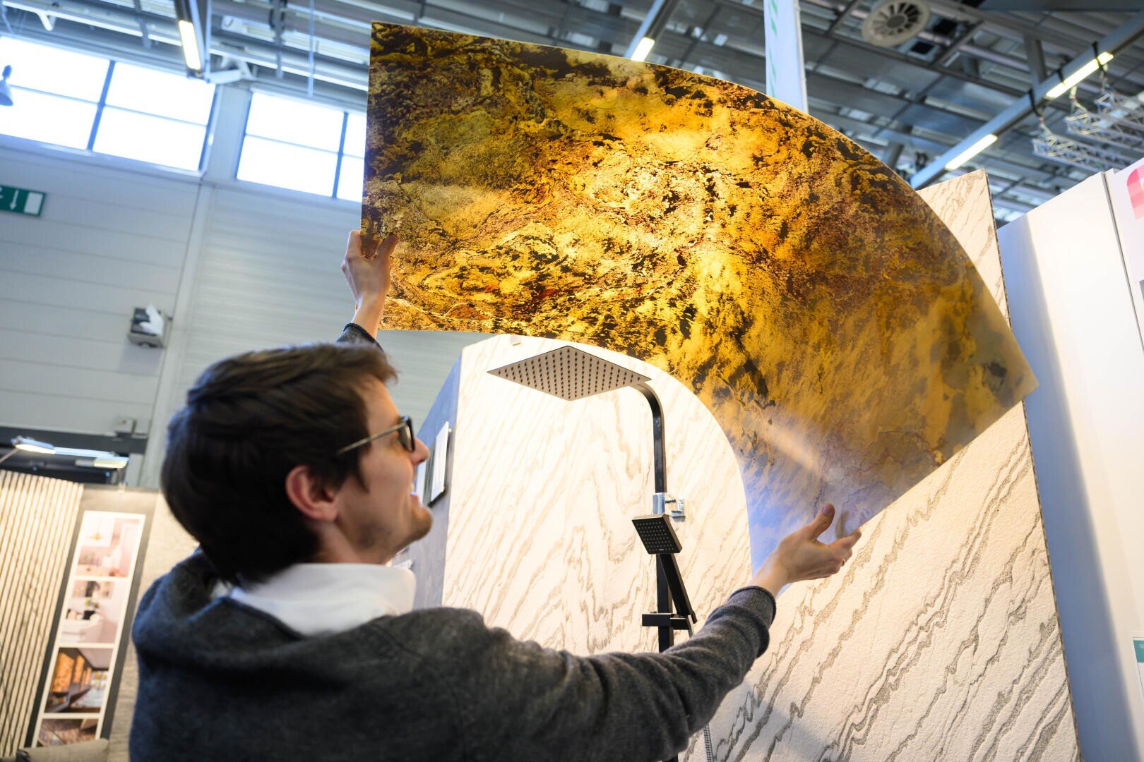 In einem modernen Ausstellungsraum hält eine Person unter hellem Licht ein großes, durchscheinendes Laken mit einem gold-braunen Marmormuster hoch.
