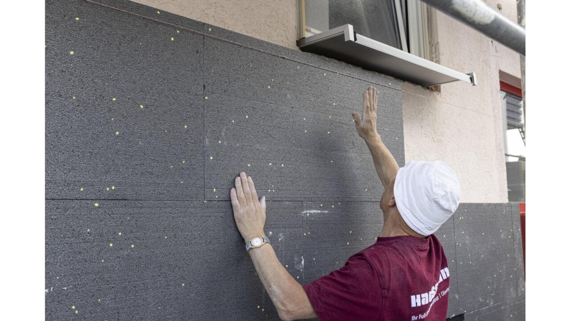 A person wearing a white hat and maroon shirt installs large gray insulation panels on an exterior wall beneath a window.