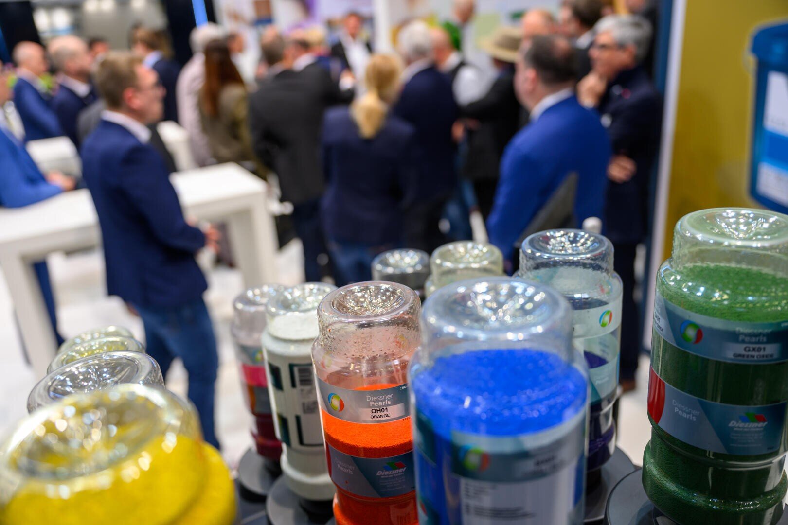 Im Vordergrund stehen auf einem Tisch Gläser mit buntem Kunststoffgranulat, während sich im unscharfen Hintergrund eine Gruppe von Menschen in Geschäftskleidung bei einer Veranstaltung in einem geschlossenen Raum versammelt und unterhält.