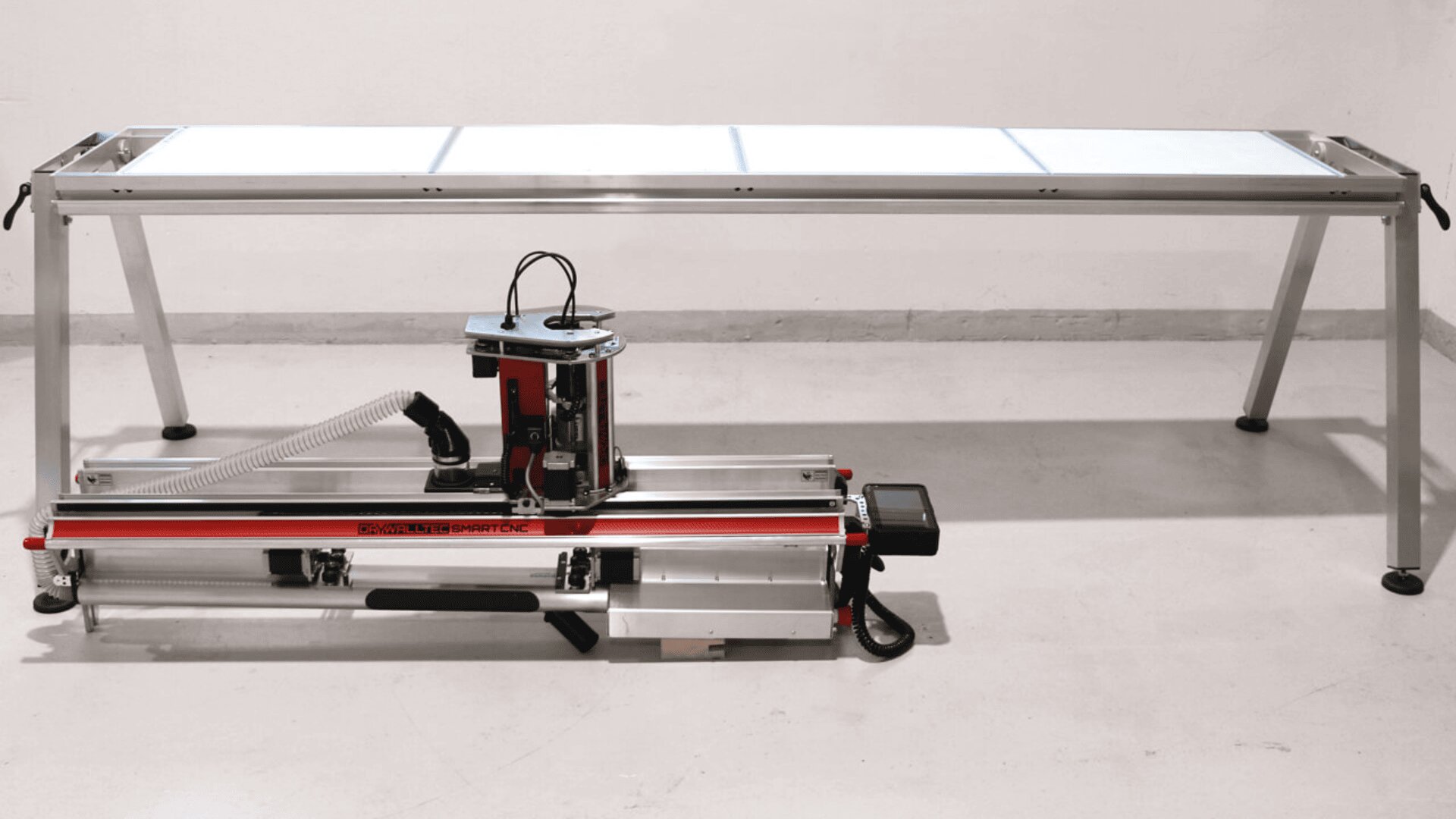 Eine CNC-Fräsmaschine mit einem rot-silbernen Gehäuse steht auf dem Boden vor einer großen rechteckigen Werkbank mit Aluminiumrahmen und weißer Oberfläche.