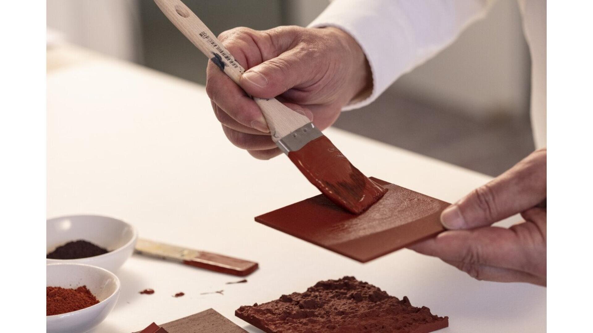 A person uses a paintbrush to apply a layer of reddish-brown paint onto a small rectangular tile, with bowls of pigment and other tiles nearby on a white surface.