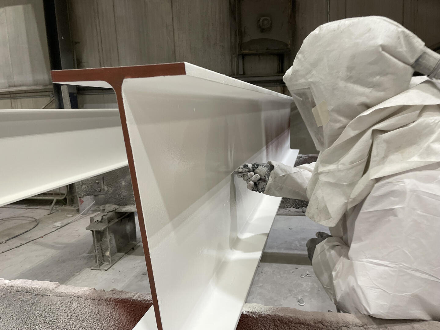 Eine Person in Schutzkleidung sprüht weiße Farbe auf einen großen Metallträger in einer Industrieanlage und achtet dabei auf eine gleichmäßige Deckung.