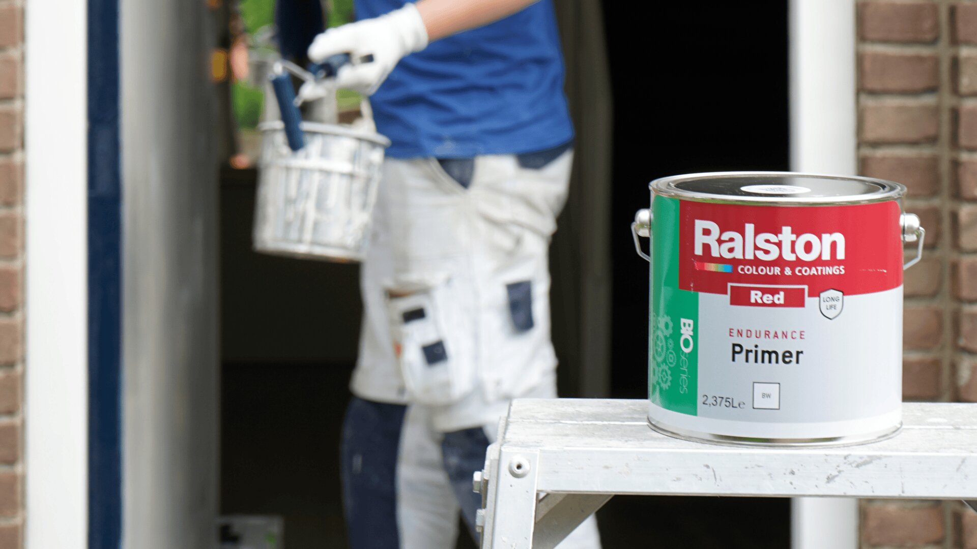 A can of Ralston red primer sits on a ladder in the foreground, while a person in painting clothes and gloves holds a paintbrush and tin, standing near a doorway in the background.