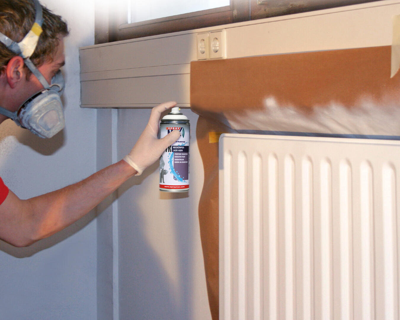 Eine Person mit Maske und Handschuhen sprüht aus einer Dose weiße Farbe auf eine Wand über einem Heizkörper, wobei braunes Schutzpapier Teile des Bereichs abdeckt.