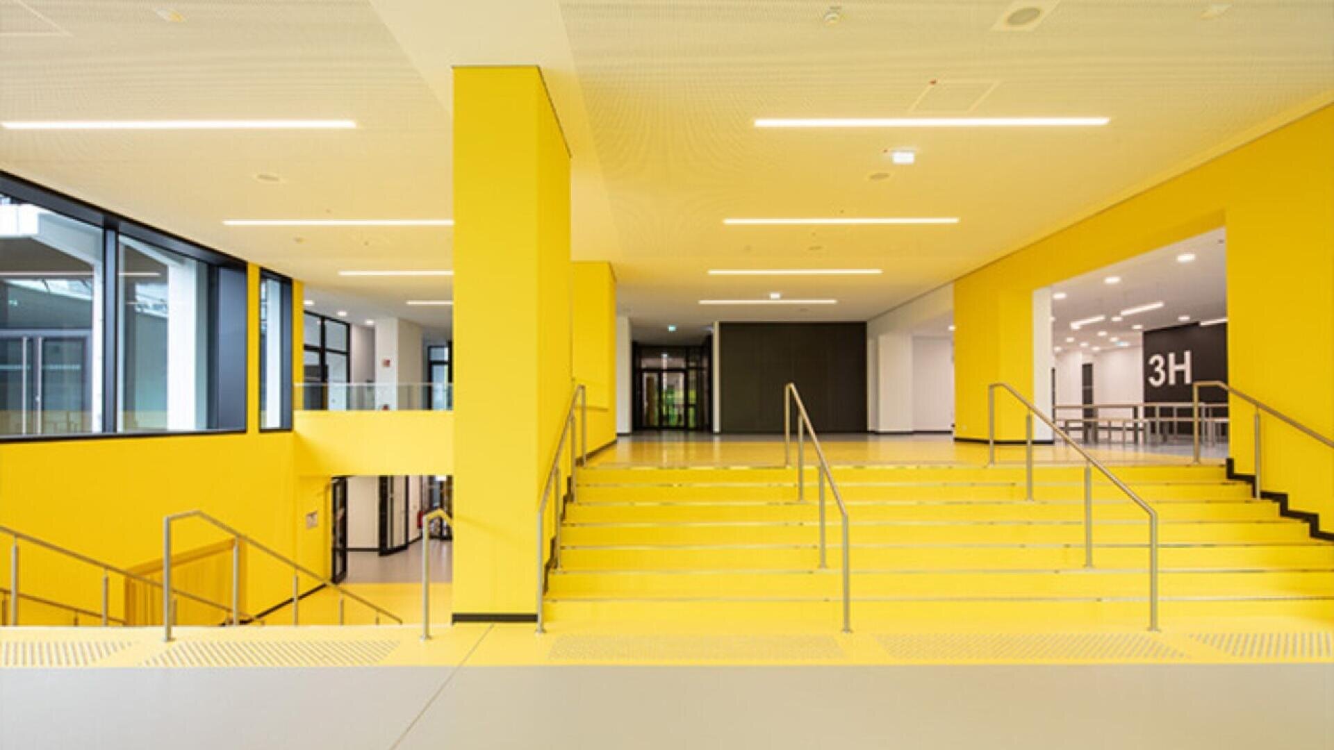 A modern, bright interior space features wide yellow stairs with metal railings, yellow walls, and large windows letting in natural light. Signs indicate room 3H in the background. The design is clean and minimalistic.