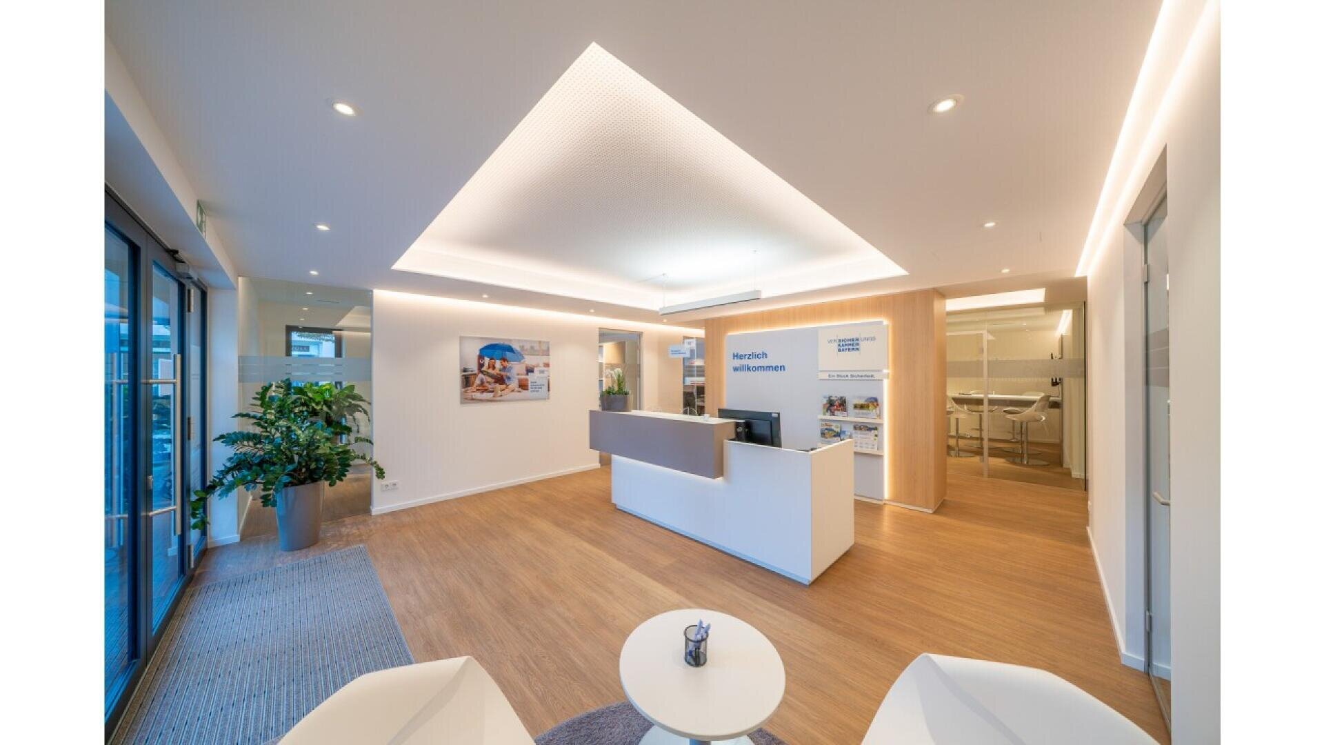 Modern office reception area with a white desk, seating, wooden flooring, large indoor plant, wall art, bright lighting, and a welcoming atmosphere.