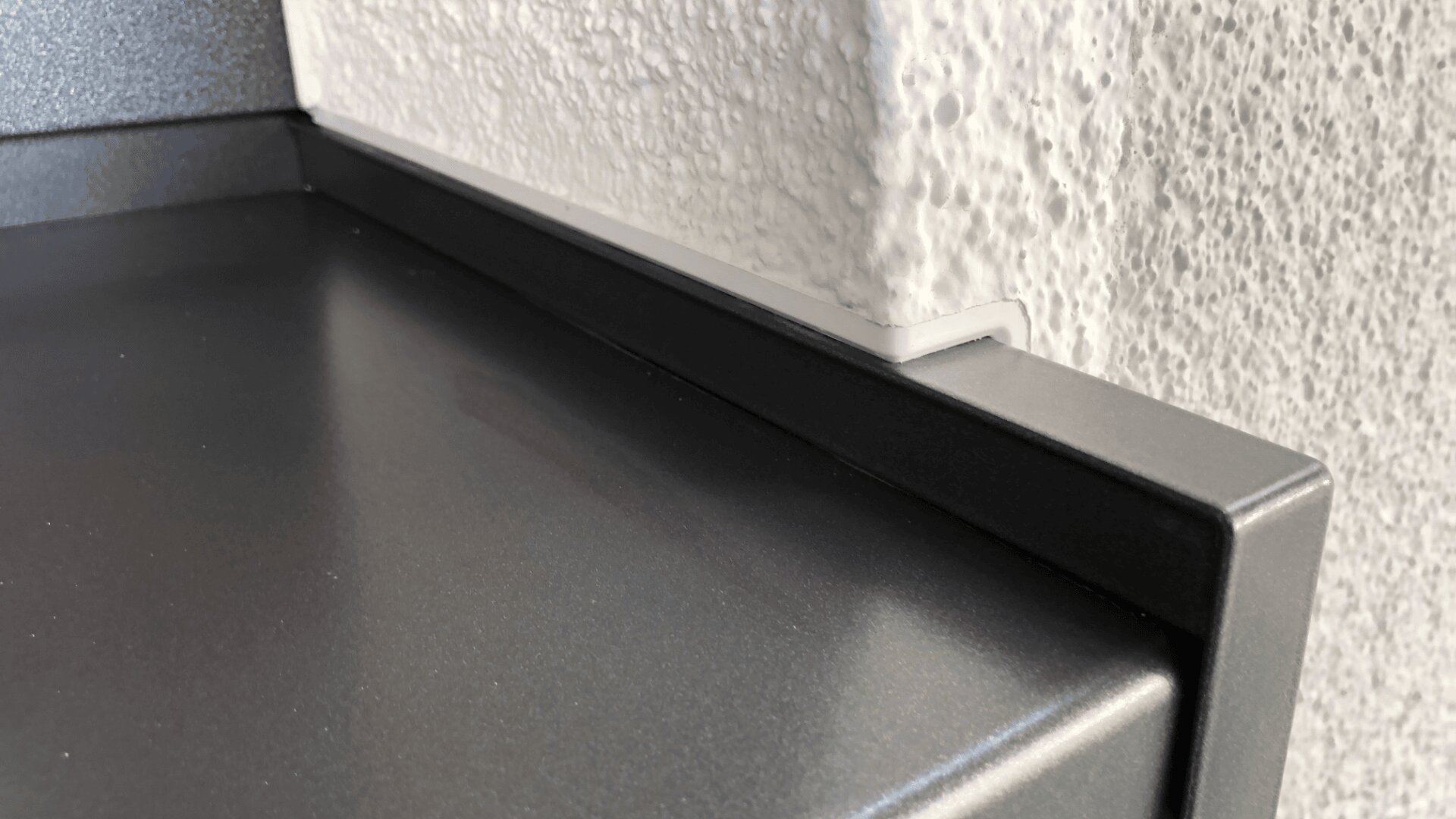 Nahaufnahme einer mattschwarzen Metalloberfläche, die eng an einer weißen strukturierten Wand anliegt, mit einem präzisen Ausschnitt in der Leiste, um einem hervorstehenden Abschnitt der Wand gerecht zu werden.
