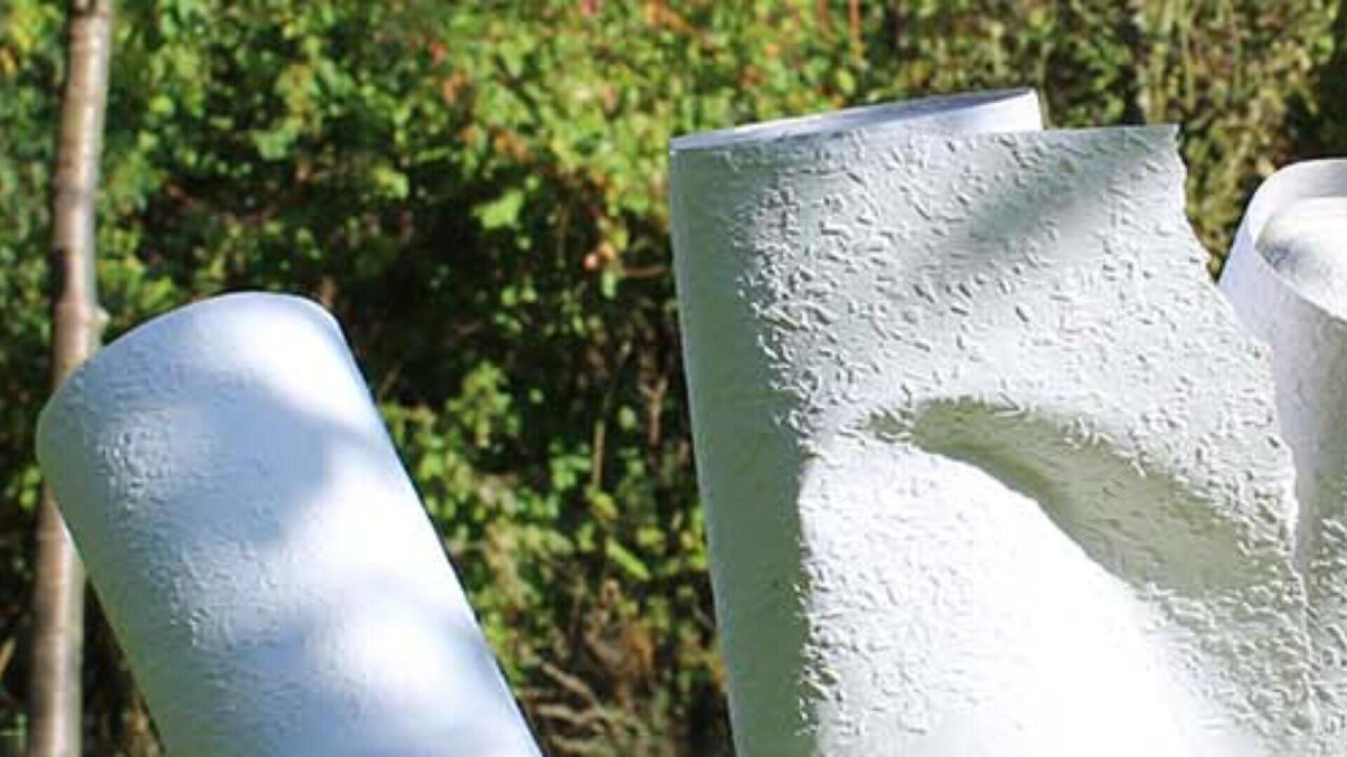 Three large, textured white cylindrical sculptures stand outdoors against a background of green trees and foliage in natural sunlight.