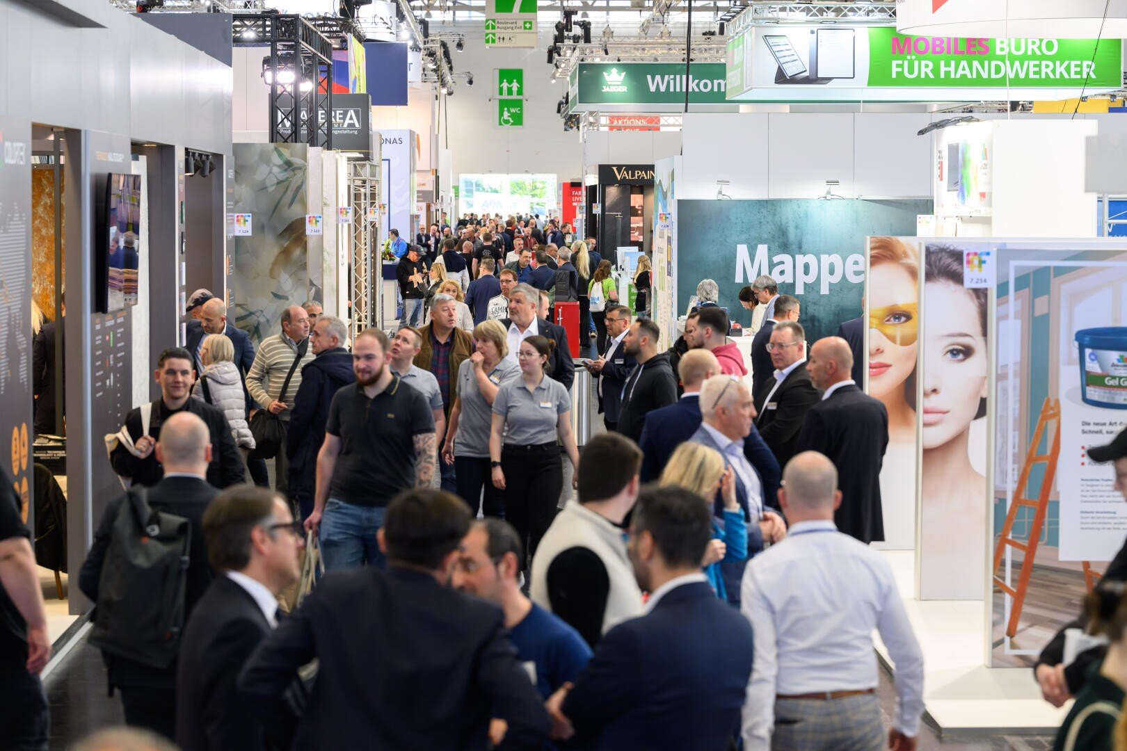 Eine geschäftige Handelsmesse mit vielen Menschen, die in einer großen Halle mit farbenfrohen Ständen, Anzeigen und Schildern unterwegs sind und sich unterhalten, darunter auch leuchtende Bilder und Banner, die über der Menge hängen.