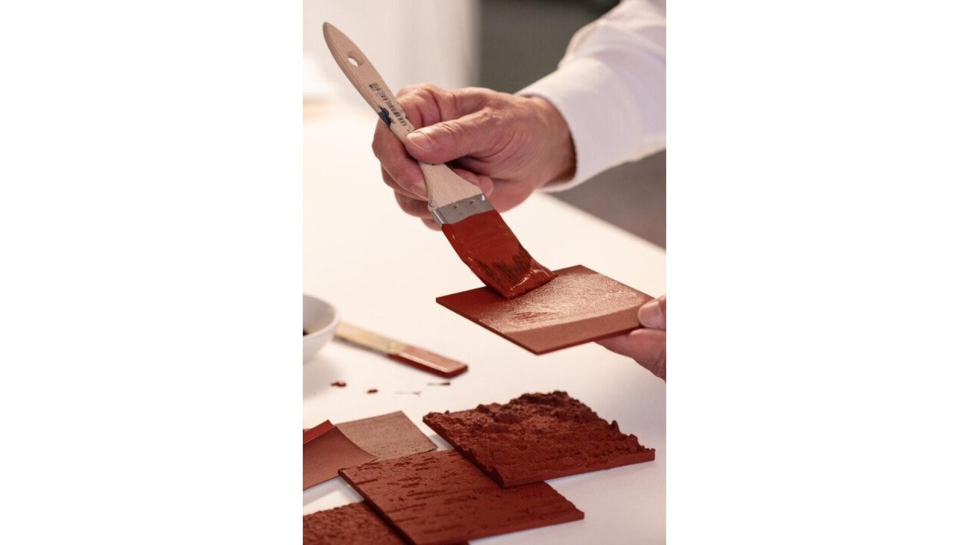 A person uses a paintbrush to apply red paint or clay onto a square tile. Several other tiles with red textured surfaces are on the table nearby.