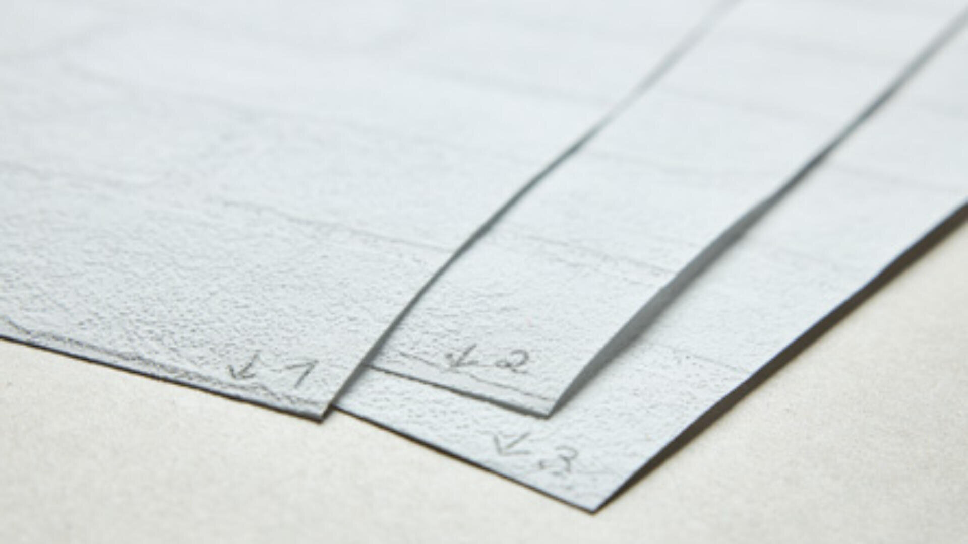 Drei übereinanderliegende Blätter strukturierten weißen Papiers sind an den Ecken mit den handschriftlichen Bleistiftzahlen „1“, „2“ und „3“ beschriftet. Die Blätter werden auf eine helle, glatte Unterlage gelegt.