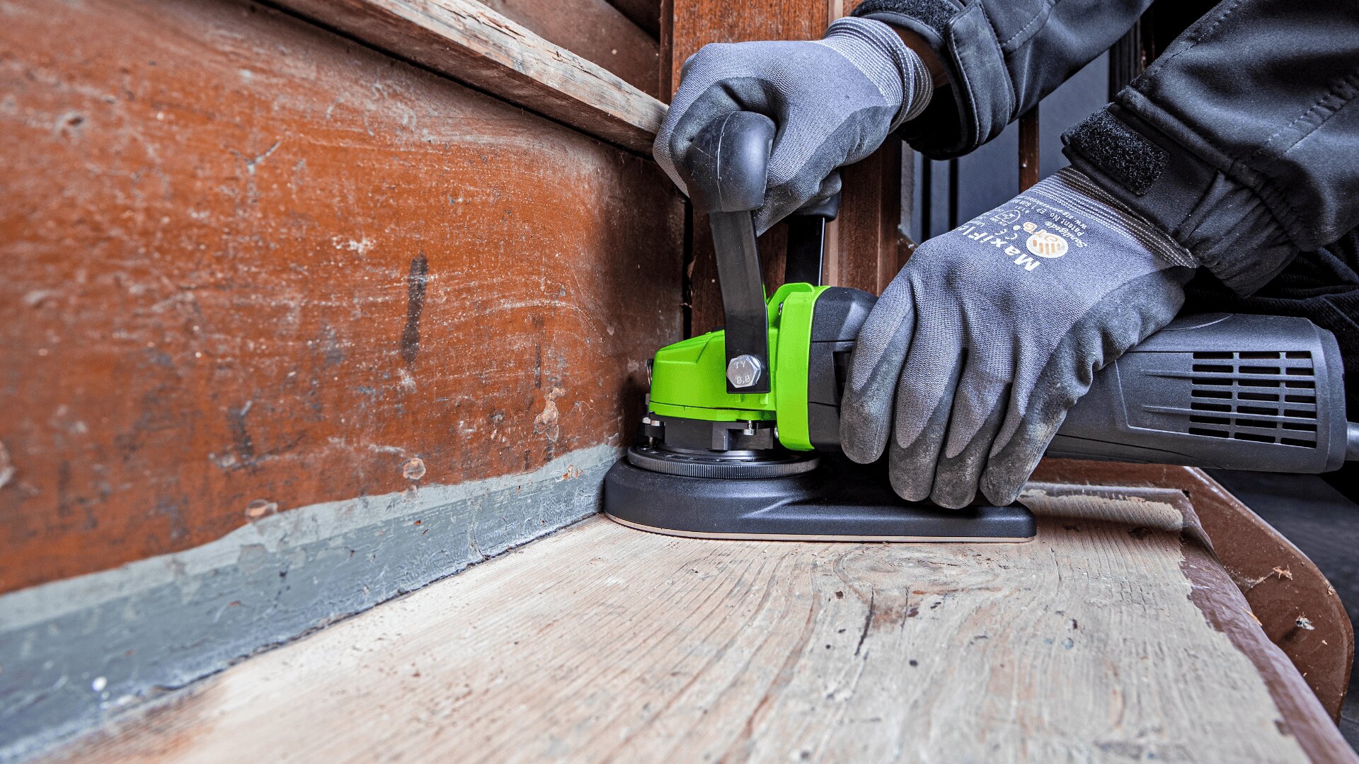 Eine Person mit Handschuhen glättet mit einem grünen elektrischen Schleifgerät die Kante, an der eine Holzwand auf einen Holzboden trifft, und konzentriert sich dabei auf detaillierte Schleifarbeiten.