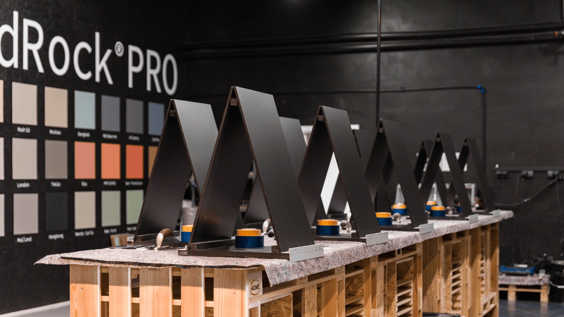 Eine Reihe schwarzer Metalldachplatten sind auf Holzpaletten in einem Ausstellungsraum ausgestellt, im Hintergrund eine Wand mit Farbmustern mit der Aufschrift „LandRock PRO“.