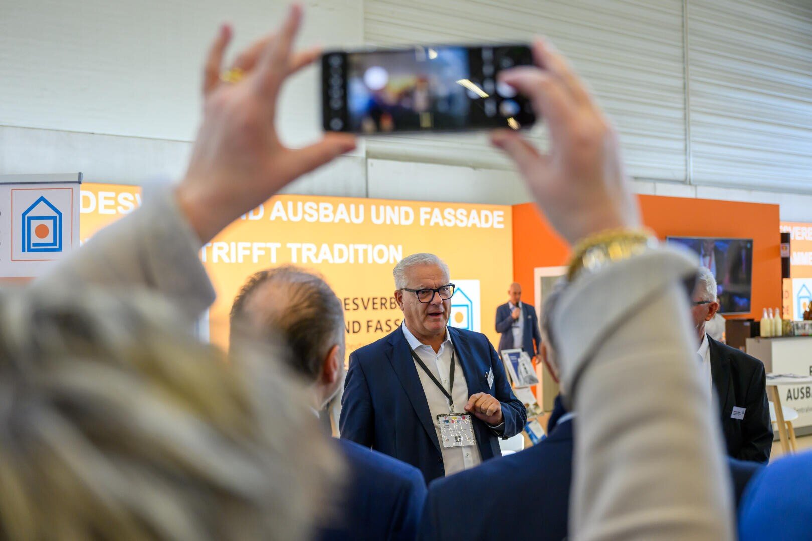 Ein Mann im Anzug spricht bei einer Indoor-Veranstaltung zu einer Gruppe, während im Vordergrund jemand ein Telefon hochhält, um ein Foto oder Video aufzunehmen. Im Hintergrund sind orangefarbene Banner mit deutschem Text zu sehen.