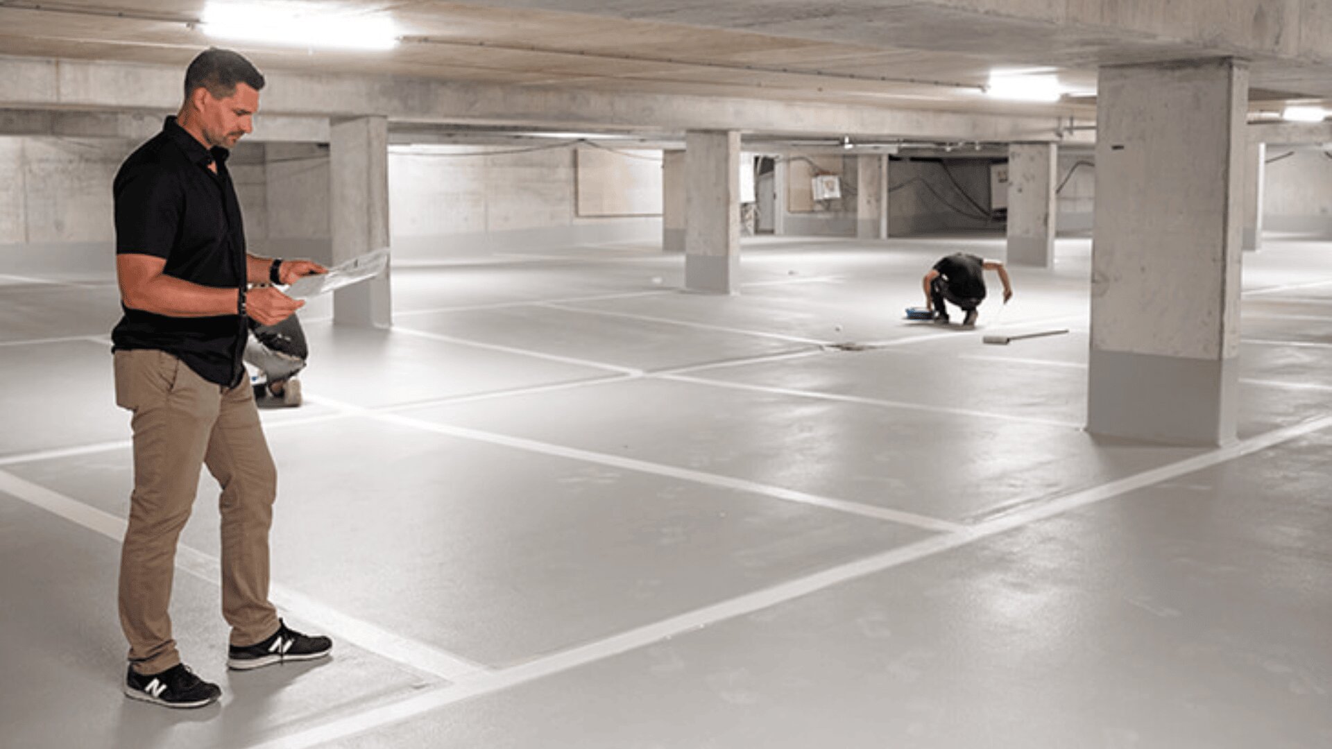 Ein Mann steht mit Papieren in einem leeren, frisch gestrichenen Parkhaus, während in der Ferne zwei Arbeiter knien und am Boden arbeiten. Die Szene ist durch Deckenleuchten gut beleuchtet.