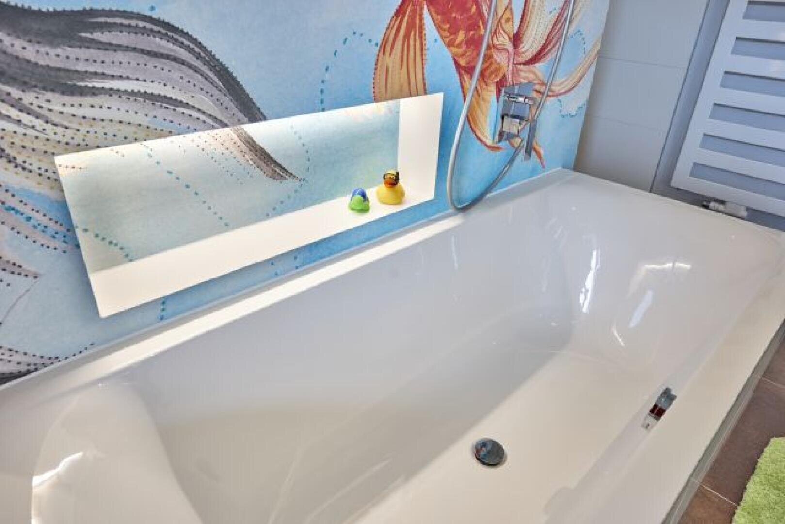 Eine weiße Badewanne unter einem farbenfrohen Wandgemälde mit Fischmotiven, mit einem modernen, beleuchteten Regal mit zwei Gummienten, einer gelben und einer grünen, neben einem an der Wand montierten Wasserhahn und Duschkopf.