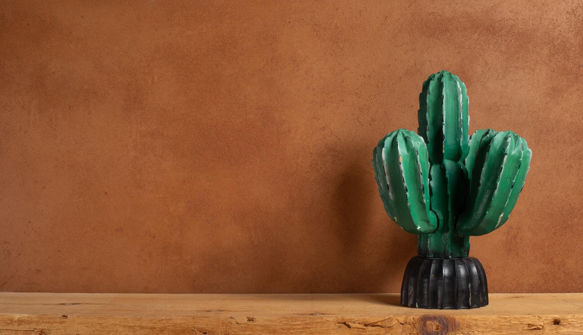 Eine grüne Kaktusskulptur in einem schwarzen Topf steht auf einer Holzoberfläche vor einer braun strukturierten Wand, mit leerem Raum auf der linken Seite.