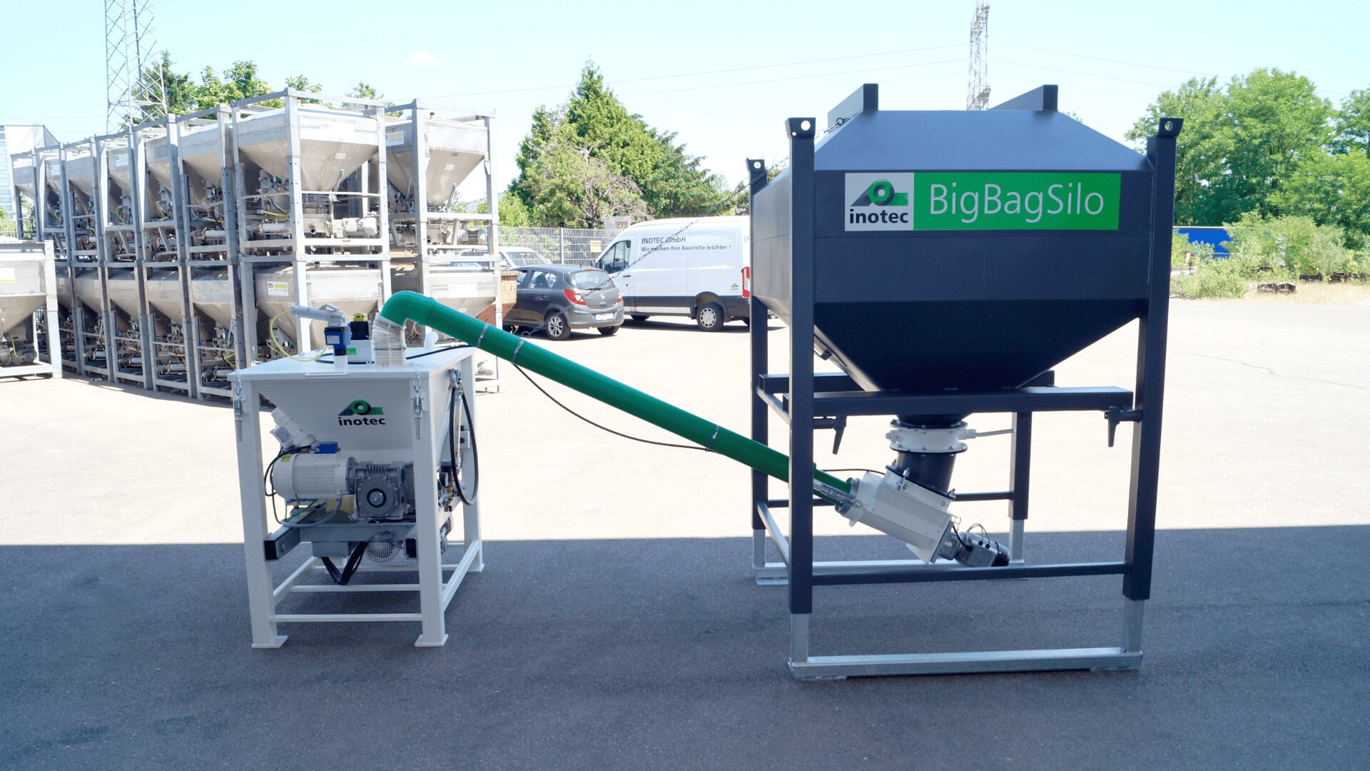 Ein großes schwarzes Silo mit der Aufschrift „BigBagSilo“ von Inotec steht im Freien neben einer weißen Steuereinheit, verbunden durch ein grünes Rohr, mit parkenden Autos, Bäumen und Lagerbehältern im Hintergrund.