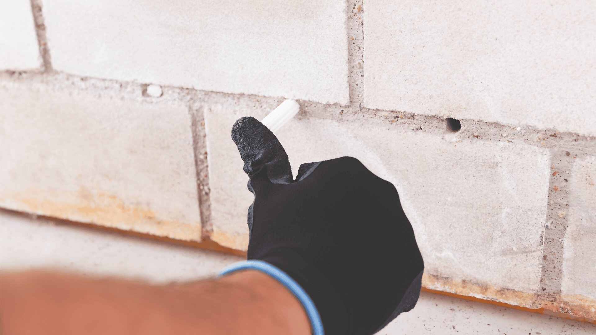 Eine Person, die einen schwarzen Handschuh trägt, führt einen weißen zylindrischen Gegenstand in ein Loch in einer weißen Ziegelwand ein.