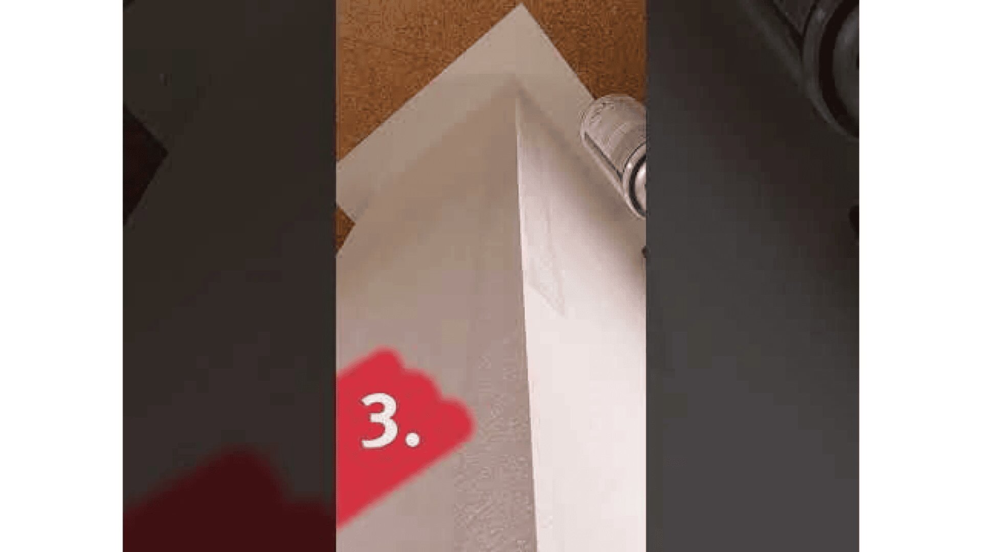 Eine Hand mit einem Tacker befestigt ein weißes Blatt Papier an der oberen Ecke einer Wand; in der unteren linken Ecke ist in Weiß auf rotem Grund die Zahl 3 zu sehen.