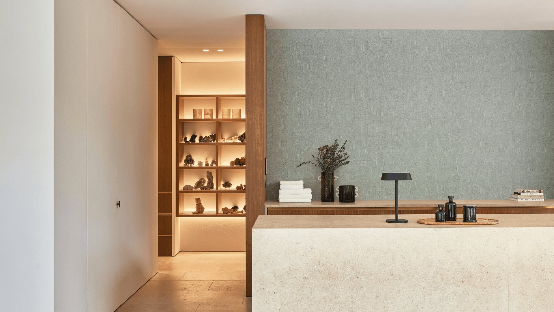 Minimalistisches Interieur mit beigefarbener Steintheke, dunkler Lampe, ordentlich gestapelten Handtüchern und Vasen. Im Hintergrund präsentiert ein sanft beleuchtetes Holzregal Deko-Objekte, teilweise von einer weißen Wand umgeben.