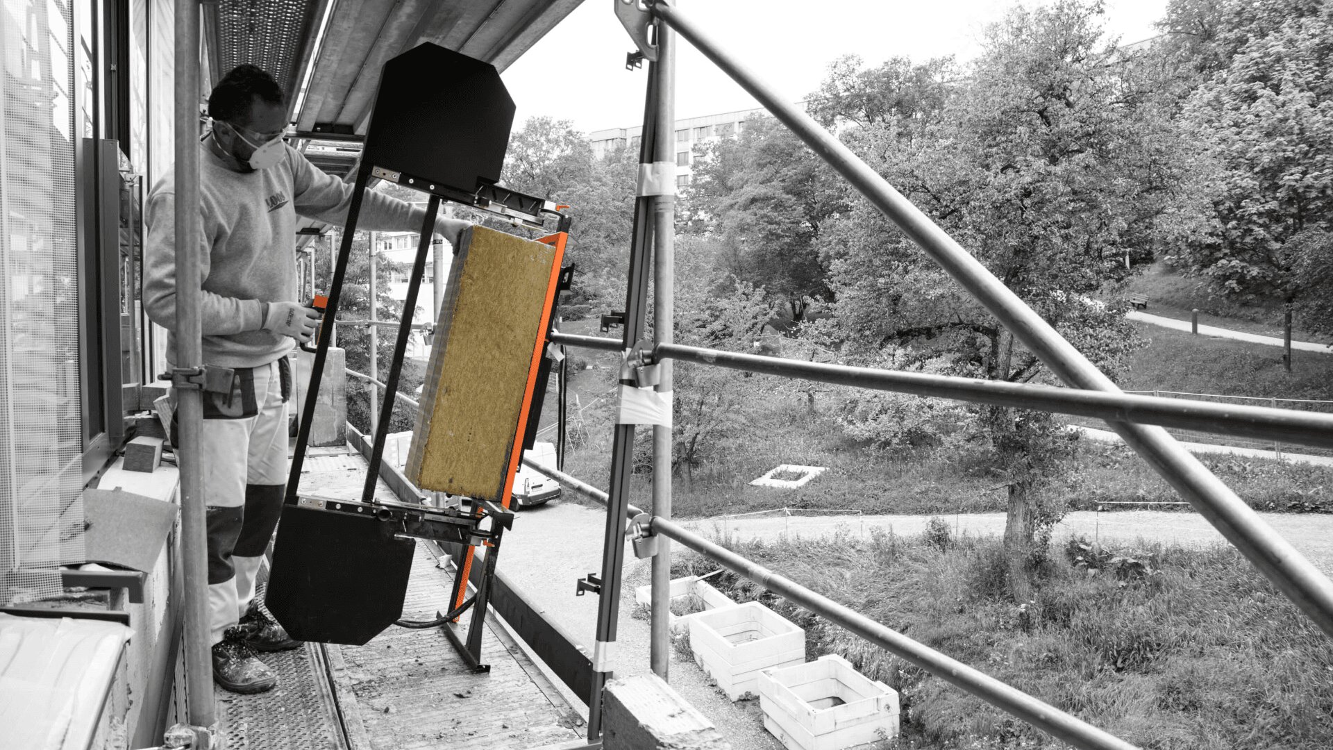 Ein Bauarbeiter auf einem Gerüst bedient Geräte neben einem Gebäude. Im Hintergrund sind Bäume und ein Weg zu sehen. Das Bild ist größtenteils schwarz-weiß, wobei die Geräte farbig hervorgehoben sind.
