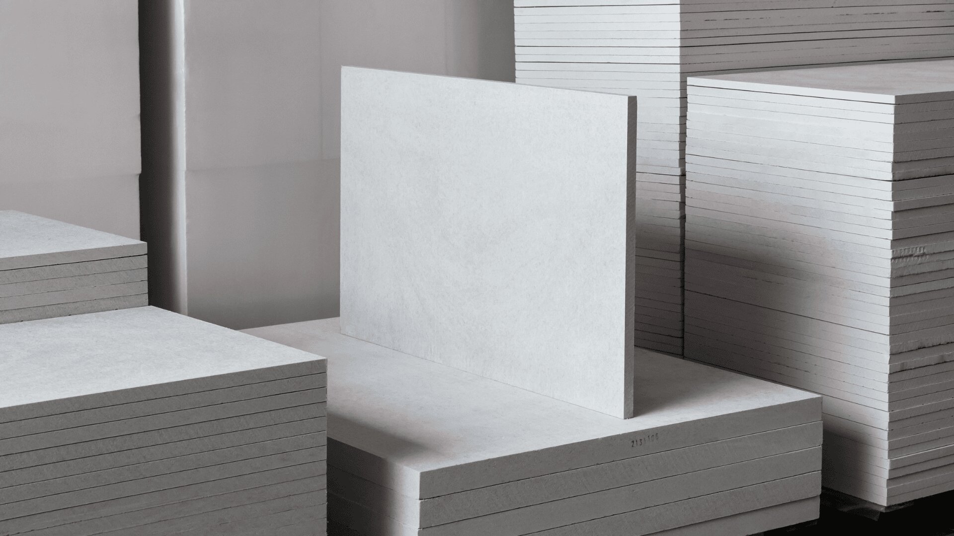 Stapel rechteckiger, hellgrauer Faserzementplatten sind ordentlich gestapelt. Eine Platte steht aufrecht an einem Stapel und zeigt ihre Oberfläche und die dünne Kante. Die Platten befinden sich in einem gut beleuchteten Innenraum.