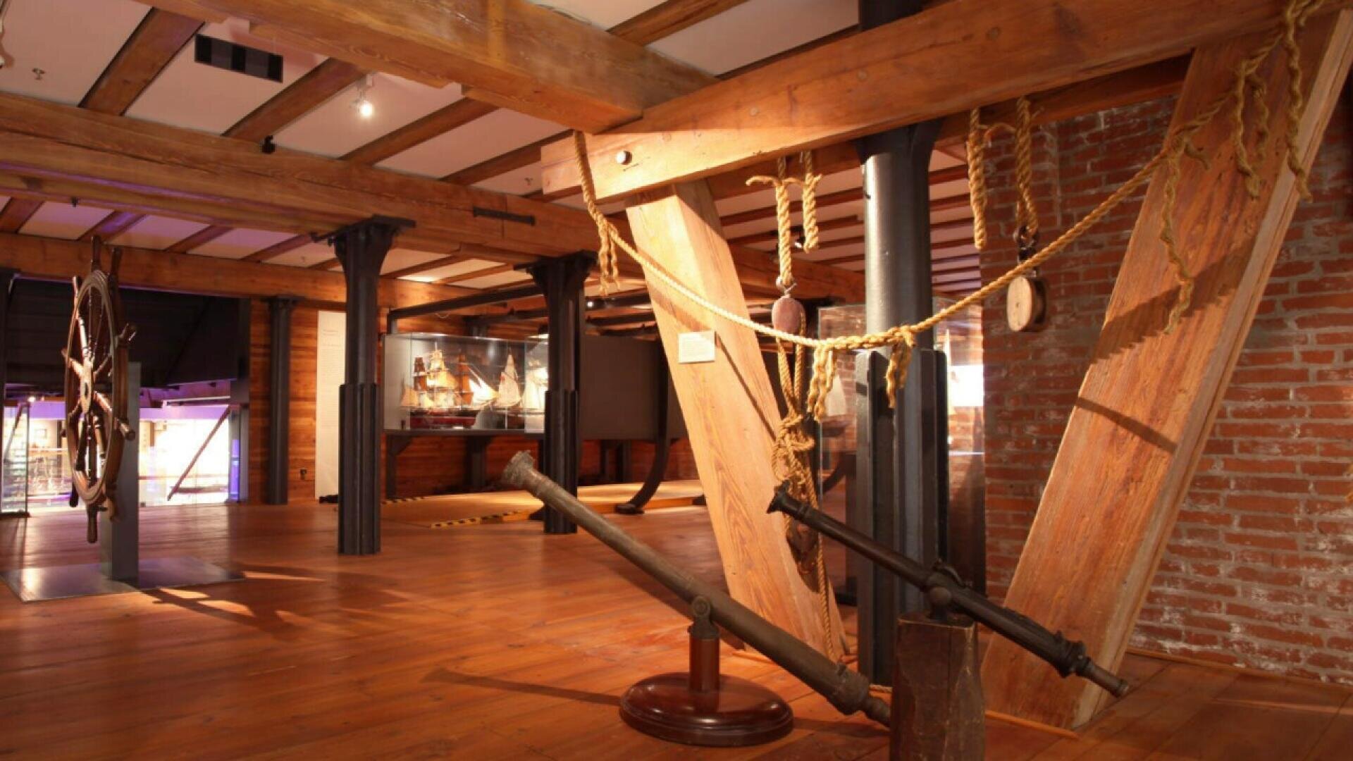 A museum exhibit with wooden beams, brick walls, and maritime artifacts including ropes and ship anchors; a glass display case with ship models is visible in the background.