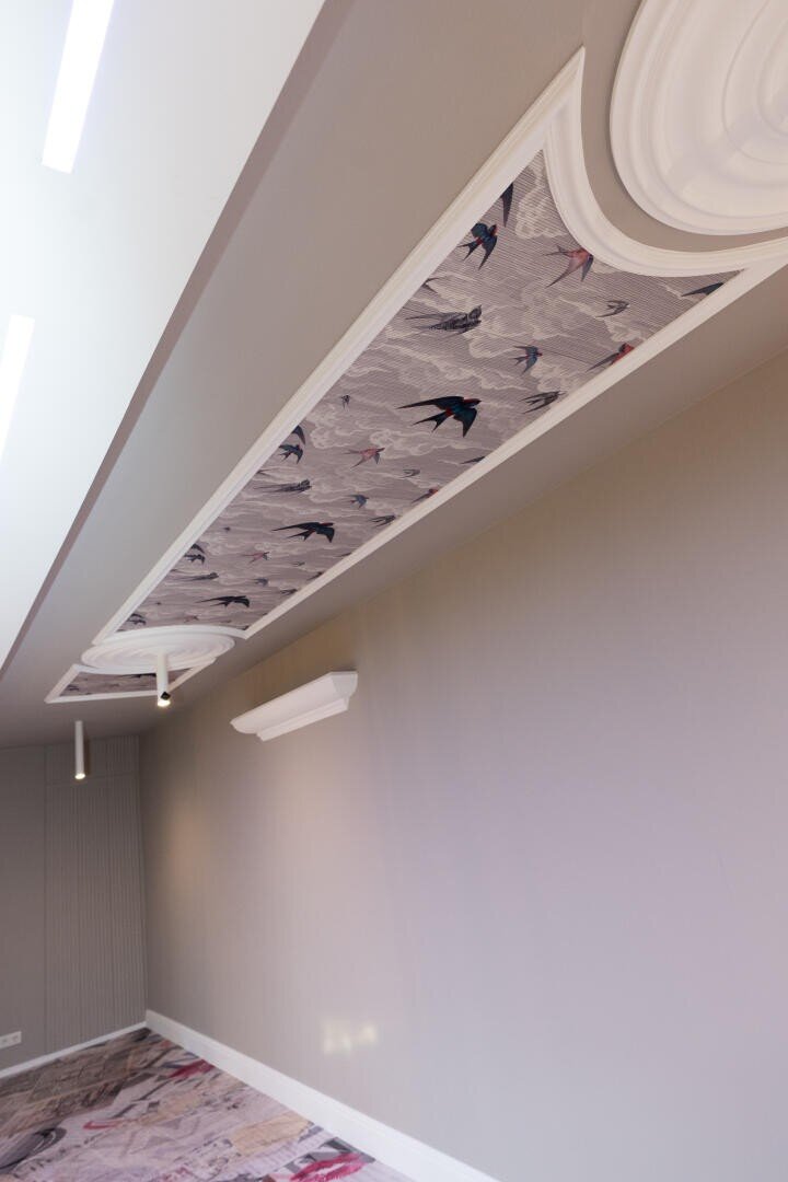 Ein verziertes Deckenpaneel mit Vogelmuster in einem modernen Raum mit hellgrauen Wänden, weißen Zierleisten und geometrischen Leuchten. Ein Teil des gemusterten Bodens ist ebenfalls sichtbar.