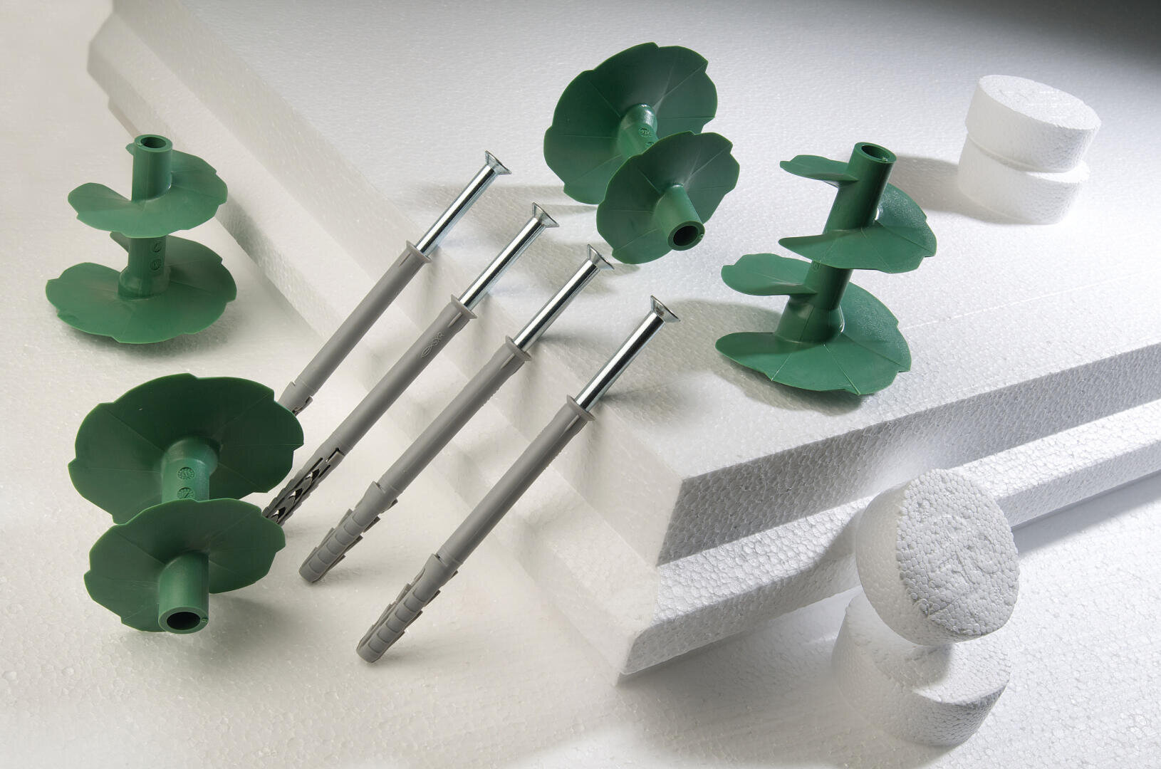 Grüne Dämmstoffbefestigungen aus Kunststoff und graue Metallschrauben sind auf und um gestapelte weiße Schaumstoffdämmplatten und zylindrische Schaumstoffstücke herum ausgestellt, die auf einer hellen Oberfläche angeordnet sind.