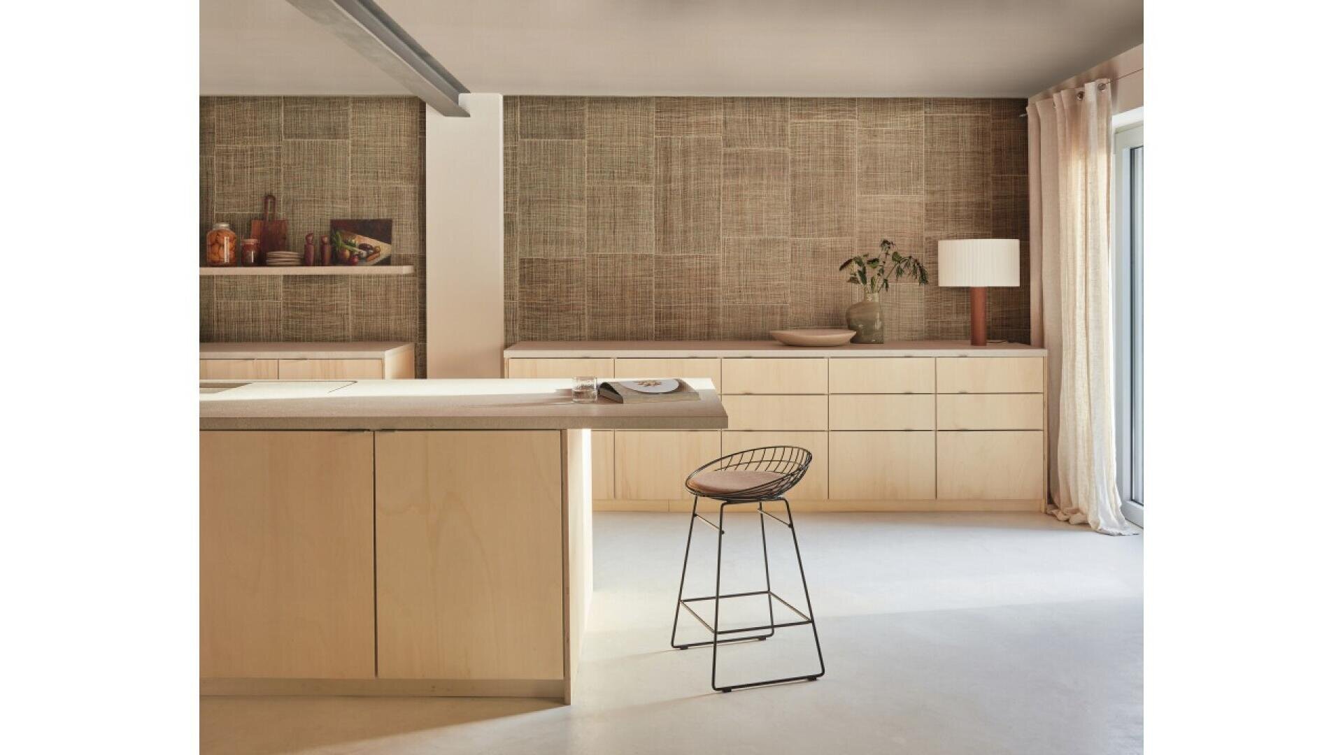 Minimalist kitchen with light wood cabinets and island, a black wire barstool, woven textured wall, bowl and lamp on the counter, and sunlight streaming through tall glass doors with light curtains.