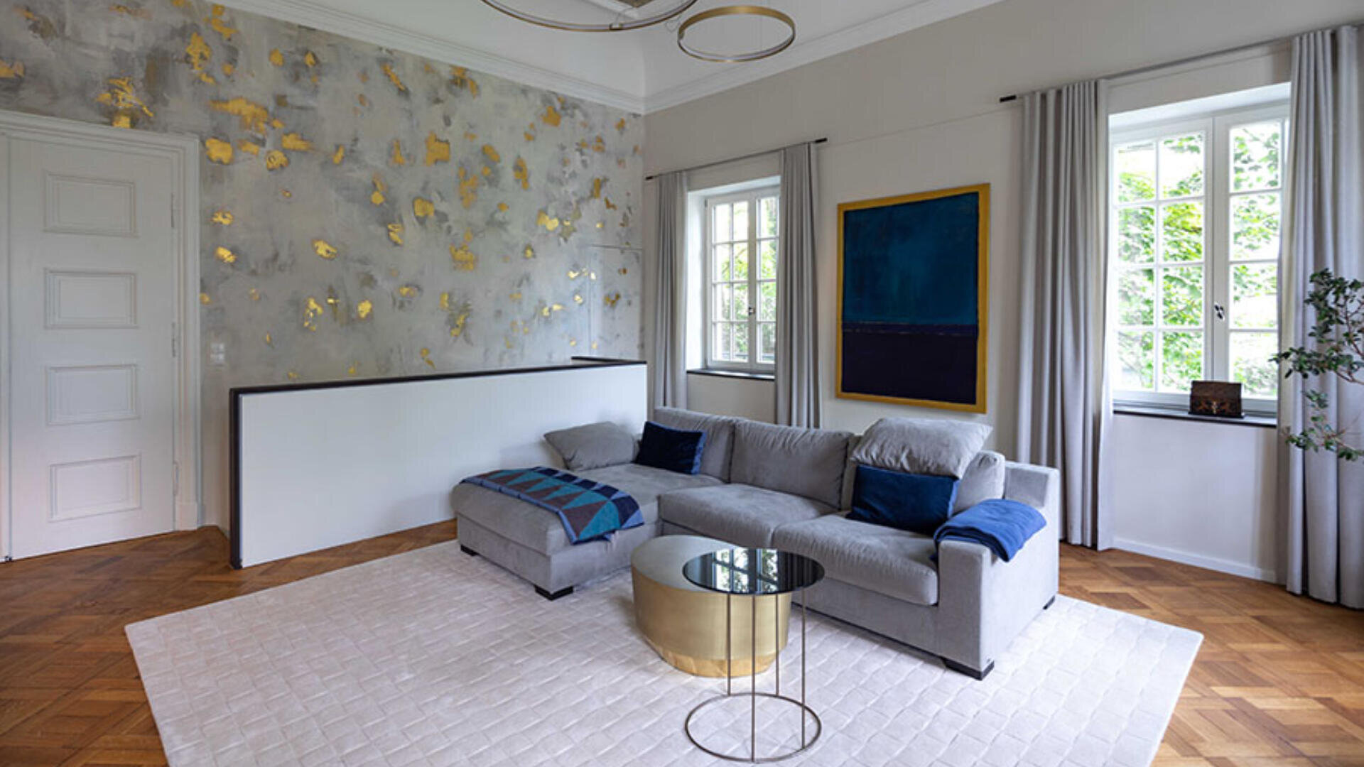 Modernes Wohnzimmer mit grauem Sofa, goldenem rundem Couchtisch, abstrakter Kunst an der Wand, großen Fenstern mit grauen Vorhängen und einem weiß gemusterten Teppich auf Holzboden. Blattgold-Akzente schmücken eine Wand.