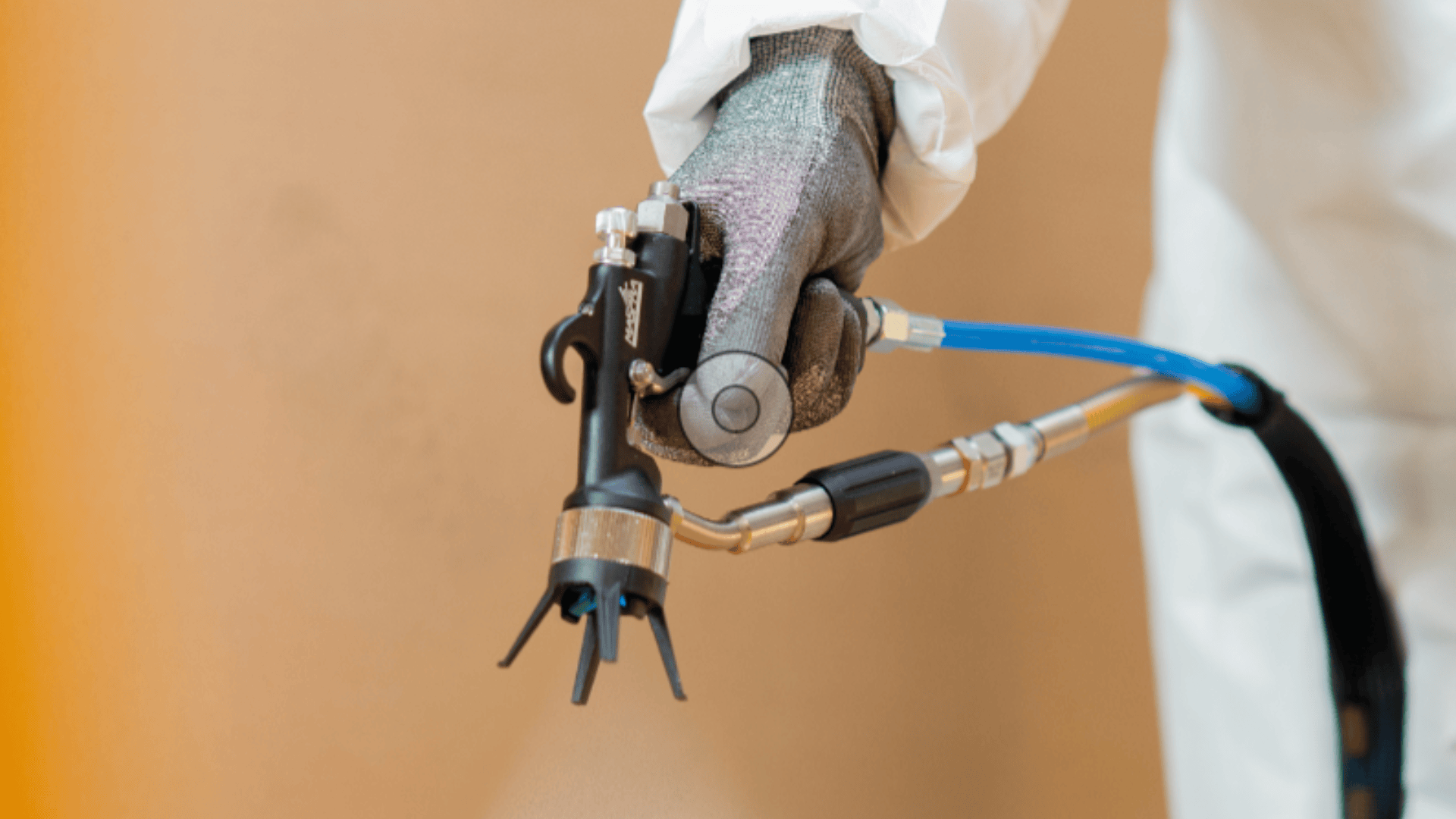 Eine Person mit Schutzhandschuhen und weißem Anzug hält vor einem schlichten Hintergrund ein spezielles Sprühgerät, das an einen blauen Schlauch angeschlossen ist und wahrscheinlich in der Industrie oder zur Schädlingsbekämpfung eingesetzt wird.